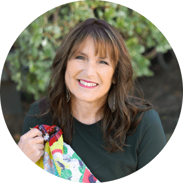 Smiling woman with brown hair and bangs, holding a colorful quilt, stands in front of green foliage. She is wearing a dark green shirt and long earrings.